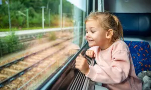 Uśmiechnięta dziewczynka patrzy przez okno pociągu. Zastanawia się, od ilu lat można jeździć samemu pociągiem, podziwiając mijane krajobrazy.