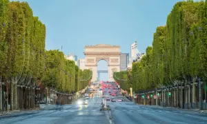 Aleja **Champs-Élysées** w Paryżu z Łukiem Triumfalnym w oddali. Drzewa tworzą zielony tunel, a ruch samochodowy płynie w stronę monumentu.