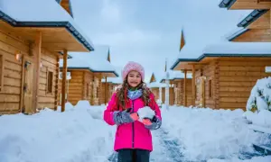 Uśmiechnięta dziewczynka w różowej kurtce trzyma kulę śnieżną. W tle drewniane domki pokryte śniegiem. Idealne ferie podkarpackie.