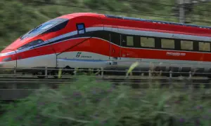 Czerwony pociąg Frecciarossa 1000 pędzi przez krajobraz, zostawiając za sobą rozmyte zielone wzgórza i kwiaty.