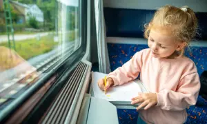 Mała dziewczynka rysuje kredką na kartce papieru, siedząc przy oknie w pociągu. Słońce oświetla jej twarz, tworząc magiczny pociąg słoneczny.