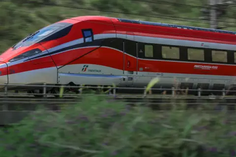 Czerwony pociąg Frecciarossa 1000 pędzi przez krajobraz, zostawiając za sobą rozmyte zielone wzgórza i kwiaty.