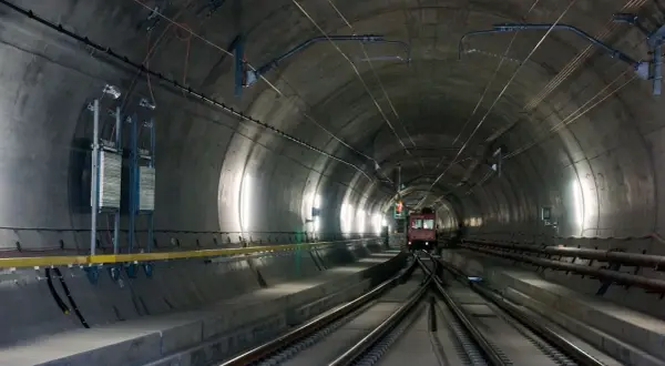 Pociąg wjeżdża do najdłuższego tunelu w Austrii. Betonowa konstrukcja, kable i oświetlenie tworzą mroczny, industrialny krajobraz.