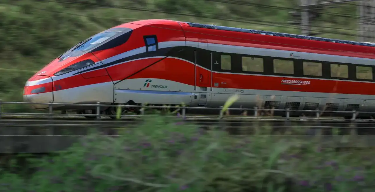 Czerwony pociąg Frecciarossa 1000 pędzi przez krajobraz, zostawiając za sobą rozmyte zielone wzgórza i kwiaty.
