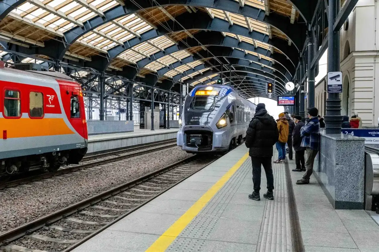 Lubuszanin pociąg na peronie, obok czerwono-pomarańczowy skład. Ludzie czekają na podróż.